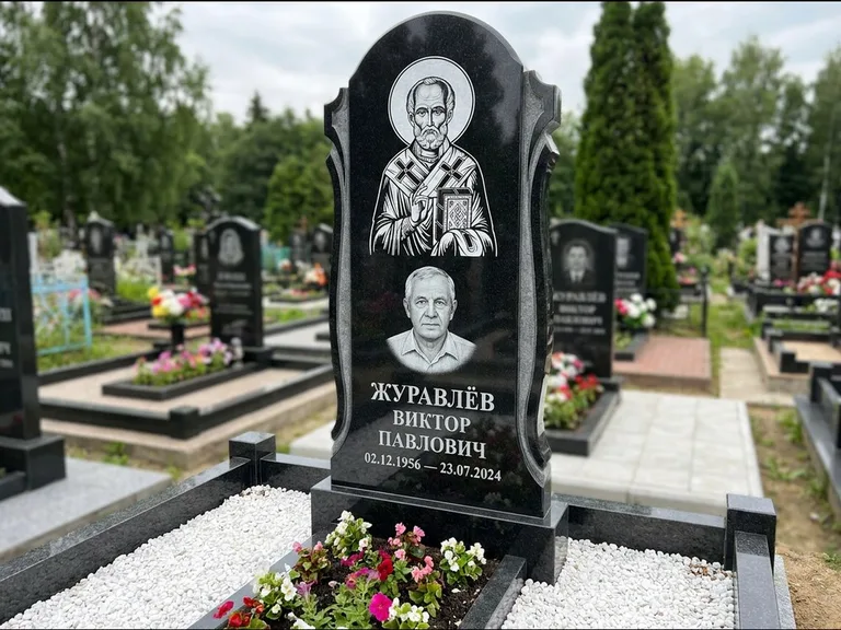 Памятник с гравировкой иконы: стела с образом в полный рост на обороте — гравировка образа Матроны Московской, низкие кусты спиреи в цвету, работа «Лебедева» в Ярославле